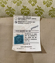 Load image into Gallery viewer, Label on a piece of furniture with text and a stamp, against a patterned fabric background.