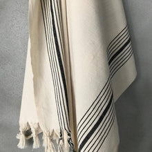 Load image into Gallery viewer, An off-white woven textile, likely a Torah shawl or prayer shawl, draped against a textured gray background. The fabric features black stripes and fringed tassels along the edge.
