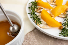 Load image into Gallery viewer, Honey dip with a spoon and crackers topped with peach slices and rosemary on a plate.