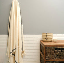 Load image into Gallery viewer, A cream-colored towel with dark gray stripes and fringed tassels hangs on a hook on a gray and white tiled wall next to a wooden side table.