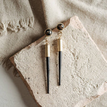Load image into Gallery viewer, Pair of earrings with gold and black components on a textured stone surface