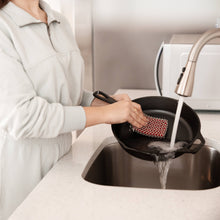 Load image into Gallery viewer, Person washing a cast iron skillet with a sponge in a kitchen sink.