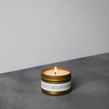 Load image into Gallery viewer, A lit gold-colored tin of Rocky Mountain Travel candle with a label that has 'ROCKY MOUNTAIN' written on it on a grey textured surface
