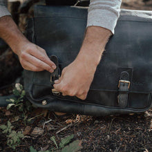 Load image into Gallery viewer, A close-up of a person's hands adjusting a metal buckle on a dark gray leather messenger bag, set against an outdoor ground background with soil and small plants.