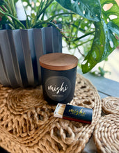 Load image into Gallery viewer, Candle with wooden lid and matchbook labeled 'mishi' on a textured surface with plants in the background.