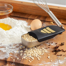 Load image into Gallery viewer, A metal measuring scoop with a black handle and three yellow silicone stripes, shown on a wooden cutting board with oats, flour, an egg, and kitchen tools.