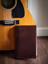Load image into Gallery viewer, Brown leather notebook leaning against a wooden acoustic guitar on a wooden surface.