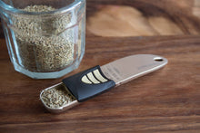 Load image into Gallery viewer, Glass container with dried herbs and a metal measuring spoon on a wooden surface
