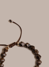 Load image into Gallery viewer, A close-up of a beaded bracelet with faceted dark brown wooden beads on a brown adjustable cord, accented with small gold spacers, set against a solid beige background.