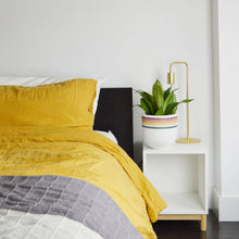 Load image into Gallery viewer, Bedroom with yellow bedding, white nightstand, and potted plant.