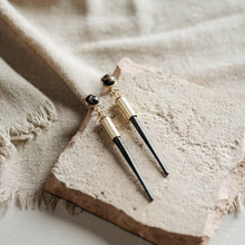 Load image into Gallery viewer, Pair of earrings with gold and black components on a textured beige surface