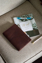 Load image into Gallery viewer, Brown leather wallet on a beige couch with a magazine and pen nearby

