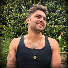 Load image into Gallery viewer, Man wearing a black tank top with tattoos and a necklace, standing in front of green foliage.
