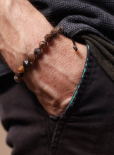 Load image into Gallery viewer, A close-up of a person's hand wearing a beaded bracelet made of brown and black faceted beads, paired with a small silver-toned ring on a black shirt and dark pants.
