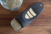 Load image into Gallery viewer, A black metal measuring cup with three stripes shown filled with dried herbs on a wooden table next to a glass jar of the same herbs.
