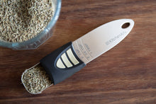Load image into Gallery viewer, Measuring spoon filled with dried herbs next to a glass container on a wooden surface
