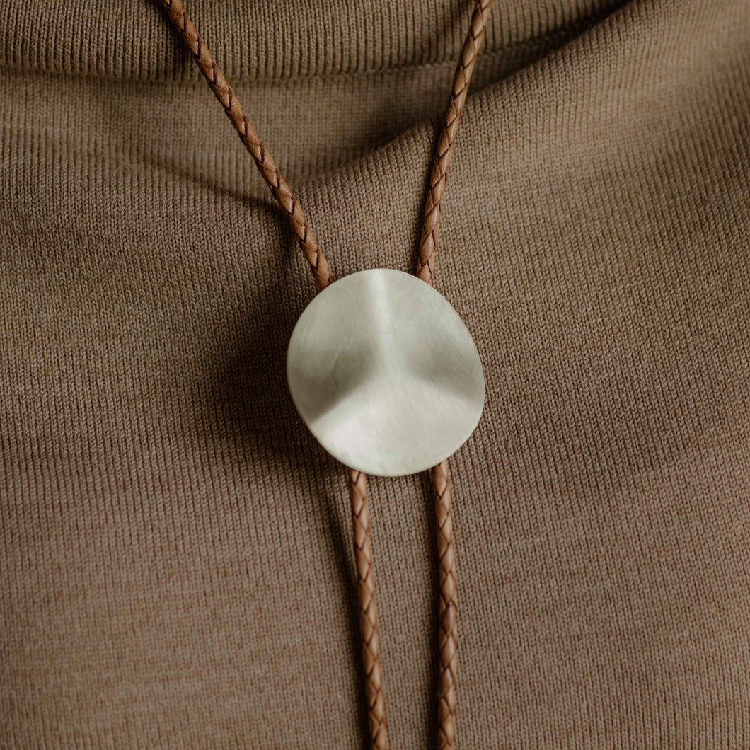 A person wearing a beige off-shoulder sweater with a brass bolo tie featuring a braided genuine leather cord.