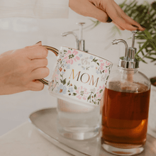 Load image into Gallery viewer, Hand holding a floral mug with 'MOM' printed on it in a coffee bar setting.
