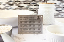 Load image into Gallery viewer, Metal measuring equivalence chart on a kitchen counter with measuring cups and containers.

