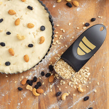 Load image into Gallery viewer, An adjustable measuring cup is shown dispensing oats and cashews onto a wooden surface next to a tart crust topped with almonds, cashews, and raisins.
