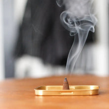 Load image into Gallery viewer, Incense cone burning on a gold tray with smoke rising against a blurred background
