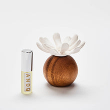 Load image into Gallery viewer, A small cylindrical perfume or diffuser bottle with a silver cap stands beside a round wooden holder topped with a white flower-shaped diffuser, photographed on a clean white background. The bottle label shows the brand ANDQ.
