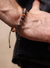 Load image into Gallery viewer, A close-up of a person's wrist and hand wearing a beaded bracelet made of faceted brown, gray, and amber stones on a black cord, with a small silver bead at the end.
