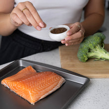 Load image into Gallery viewer, Person seasoning salmon fillet with spices on a kitchen counter.
