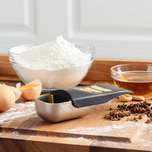 Load image into Gallery viewer, A metal measuring scoop with a black handle and yellow measurement markings, placed on a wooden cutting board with baking ingredients including flour, eggs, and spices around it.
