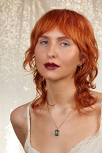 Load image into Gallery viewer, A close-up portrait of a woman with vibrant red hair, wearing a gold necklace with a rectangular blue-green pendant, a white lace camisole, and gold hoop earrings, set against a sparkly white background.
