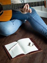Load image into Gallery viewer, Person playing an acoustic guitar with a notebook and pen on a wooden floor.

