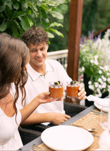 Load image into Gallery viewer, Two people sitting at a table outdoors, holding small cups.
