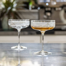 Load image into Gallery viewer, Two crystal glasses, one filled with a golden liquid, on a reflective surface.
