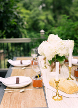 Load image into Gallery viewer, Elegant outdoor dining table setting with white flowers, gold candle holders, and greenery in the background.
