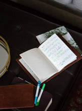 Load image into Gallery viewer, Open notebook with handwritten notes on a dark surface, accompanied by markers.
