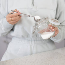 Load image into Gallery viewer, Person holding a glass jar with a white lid and a spoon, filled with a white powdery substance.

