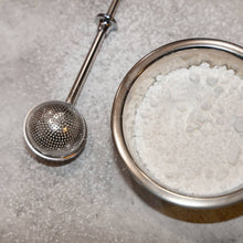 Load image into Gallery viewer, Metallic mesh strainer with a ladle over a bowl of white granulated material on a gray surface.
