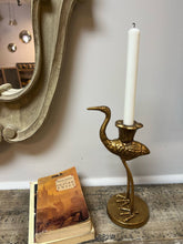 Load image into Gallery viewer, Gold crane candle holder with a white candle on a surface next to a book, with a mirror and light fixture in the background.
