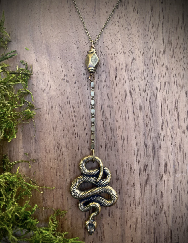 Solid 14 inch oxidized brass chain features a 4 inch charm and chain drop to a wriggling serpent charm. Displayed on wood with greenery.