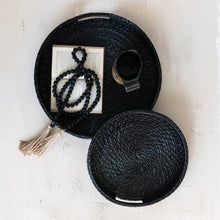 Load image into Gallery viewer, Black woven basket with a tassel, book, and cup on a textured surface
