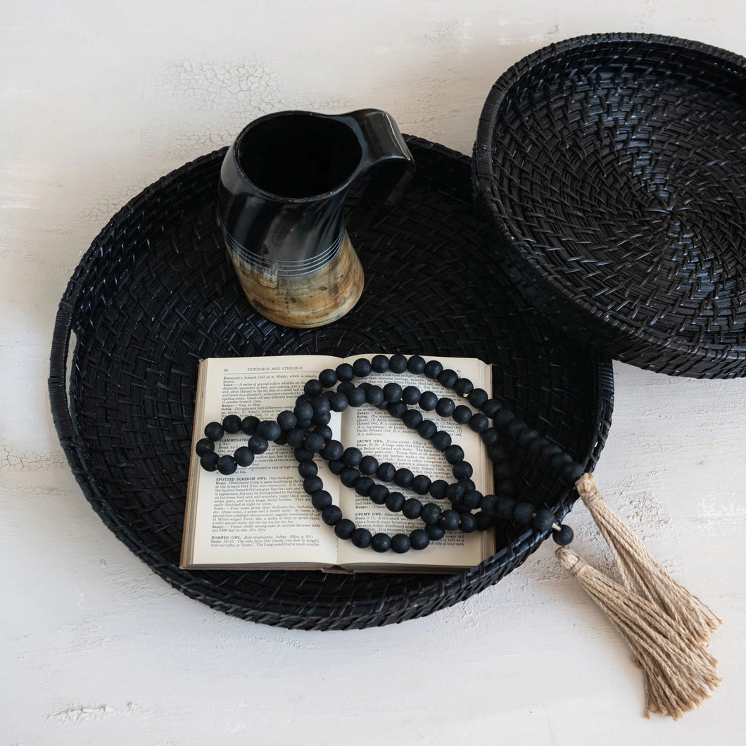 Black woven tray with an open book, black beads, and a black pitcher on a light surface.