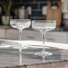 Load image into Gallery viewer, Two clear glass champagne coupes on a marble surface with a blurred kitchen background.
