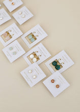 Load image into Gallery viewer, Collection of earrings on white cards with gold stands against a beige background
