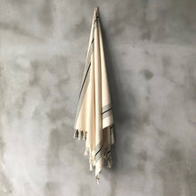 Load image into Gallery viewer, A cream-colored  towel with black stripes and tassels hangs from a hook on a textured gray concrete wall.
