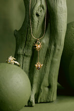 Load image into Gallery viewer, A gold pendant necklace and matching ring displayed on a textured green surface with a rustic wood prop. The jewelry features sunburst motifs with small white stones and amber accents.

