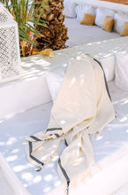 Load image into Gallery viewer, A cream-colored cotton throw blanket with black stripes and fringed edges draped over a white outdoor lounge chair, surrounded by cushions, a palm tree trunk, and a white lantern.
