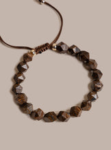 Load image into Gallery viewer, A beaded bracelet made of faceted brown gemstone beads on a dark brown adjustable cord, with small metallic spacer beads, shown on a solid beige background.
