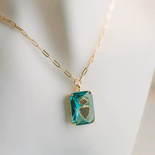 Load image into Gallery viewer, A gold chain necklace with a rectangular teal-blue crystal pendant featuring a gold heart-shaped engraving, displayed on a white mannequin bust.
