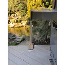 Load image into Gallery viewer, Barbecue grill with a wooden grill brush on wooden deck by a body of water and trees.
