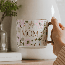 Load image into Gallery viewer, Hand holding a floral mug with 'MOM' printed on it, placed on a wooden surface.
